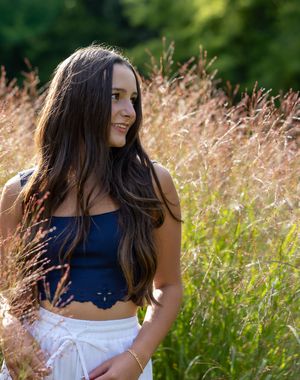 A person in a navy blue top and white skirt poses gracefully in a sunlit meadow with tall golden grass.