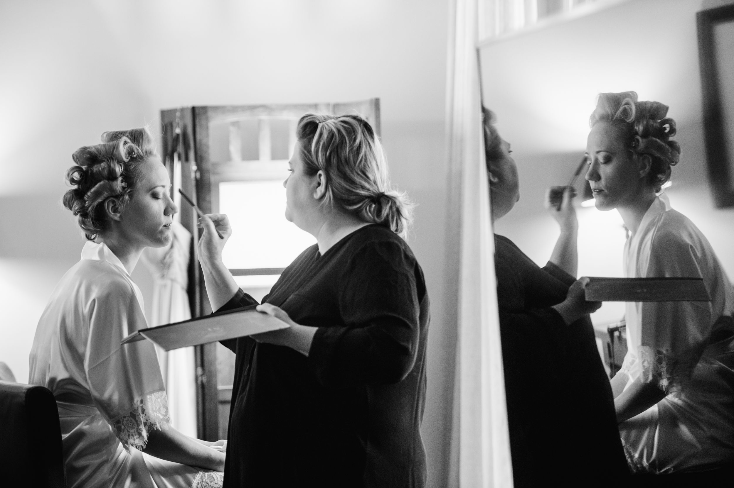A black and white photograph of figures getting ready backstage with hair rollers and costuming.
