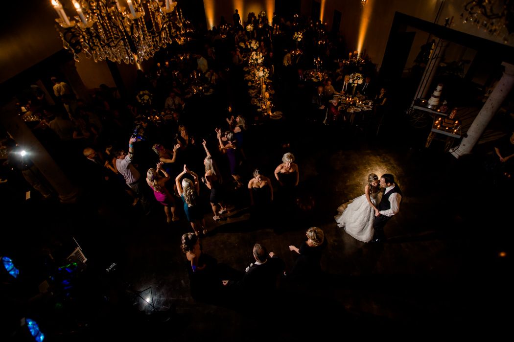 Wedding guests surround a couple dancing on the ballroom floor under elegant chandeliers at a dark reception venue.