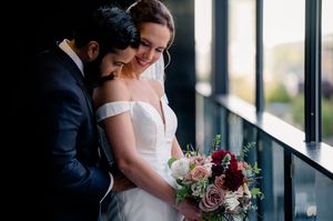 Couple embraces near large windows during intimate indoor wedding ceremony with colorful bouquet details.
