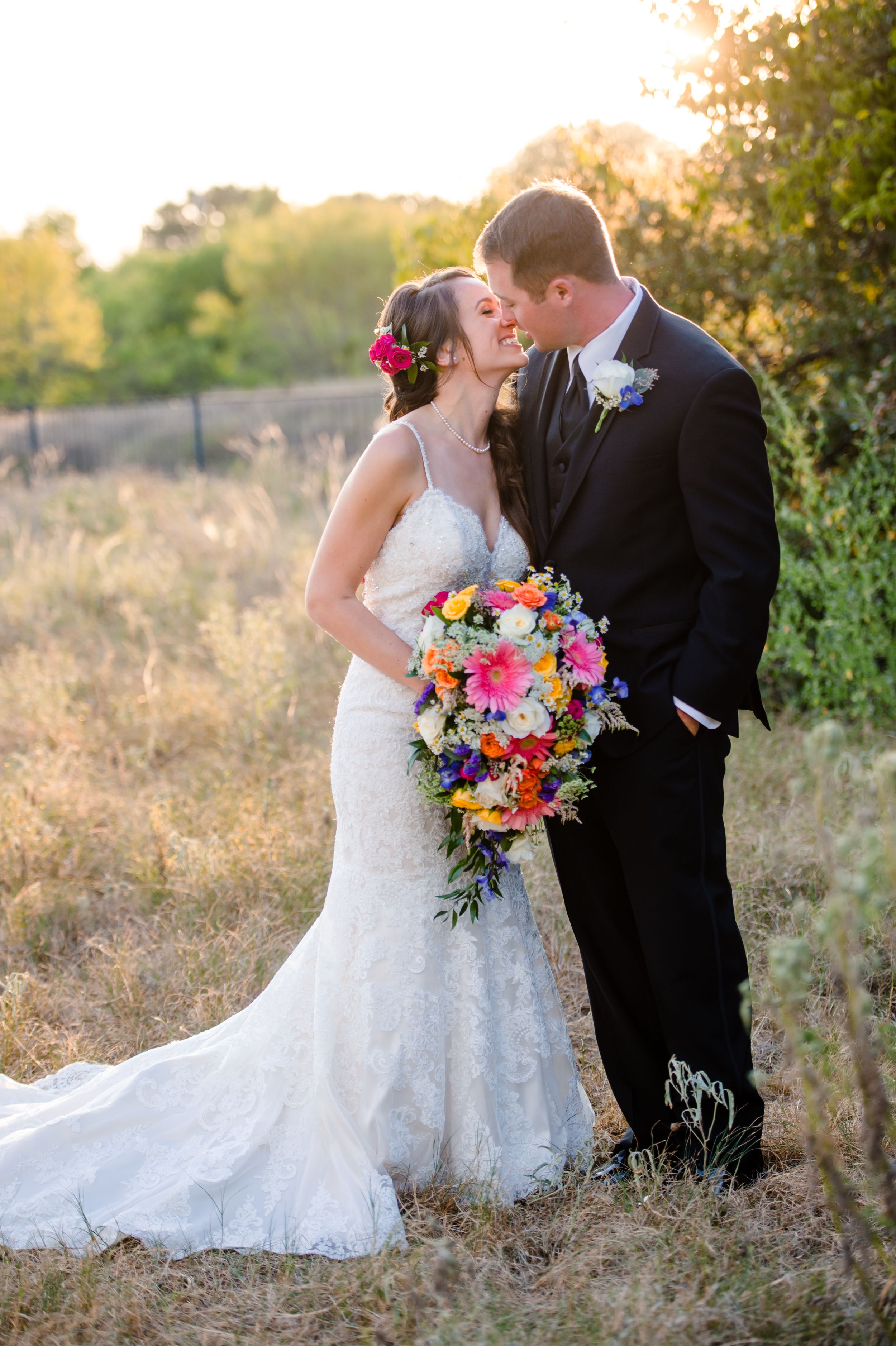 A newlywed couple shares a romantic moment at sunset in a field with the bride holding a vibrant bouquet of flowers.