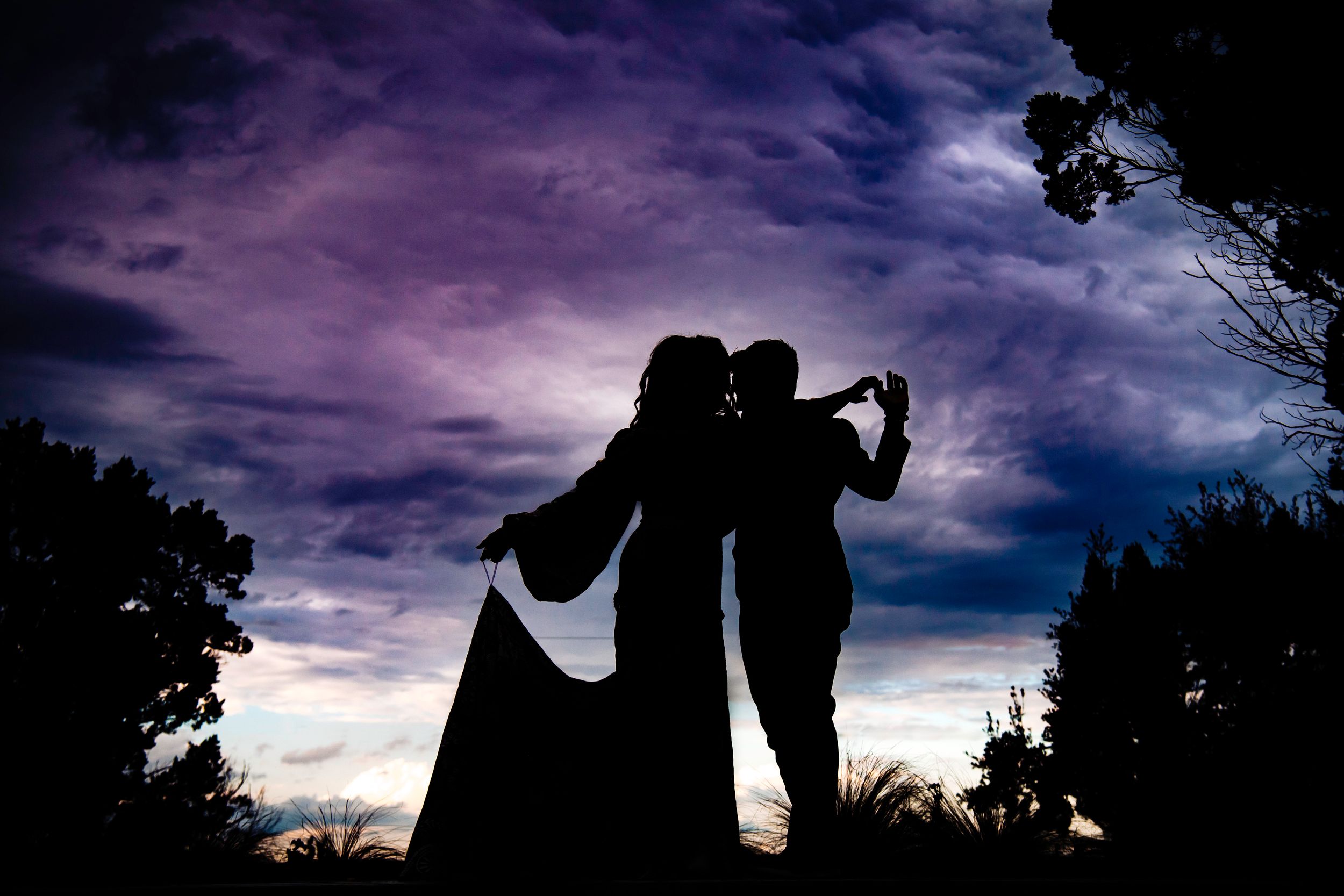 Silhouetted couple embracing against a dramatic purple and blue evening sky with trees framing the romantic scene.