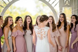 Bridesmaids in flowing pink and mauve dresses share joyful smiles and laughter with bride in white lace gown at outdoor venue.