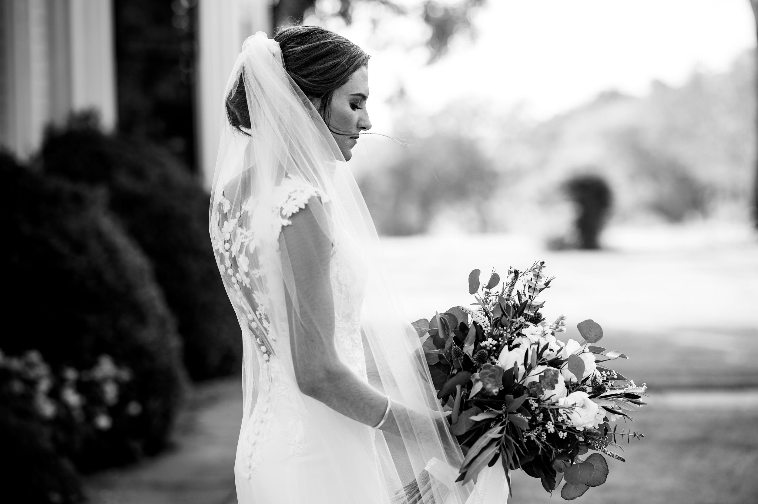 A bride in a white wedding dress holds a beautiful dark purple and white floral bouquet outdoors.