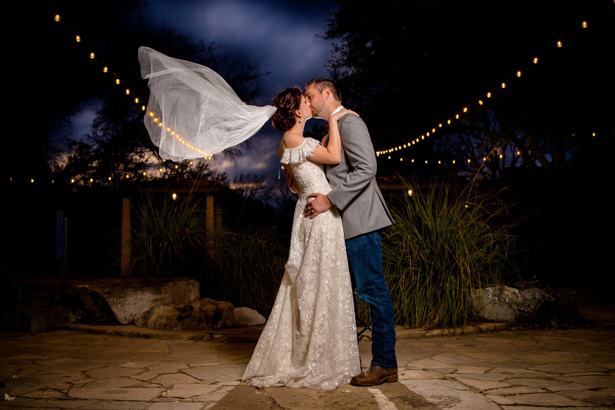 A romantic outdoor wedding scene with string lights overhead and a couple embracing on a stone patio at dusk.