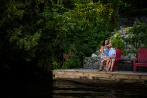 Two people sitting close together on red chairs on a dock by a lake.