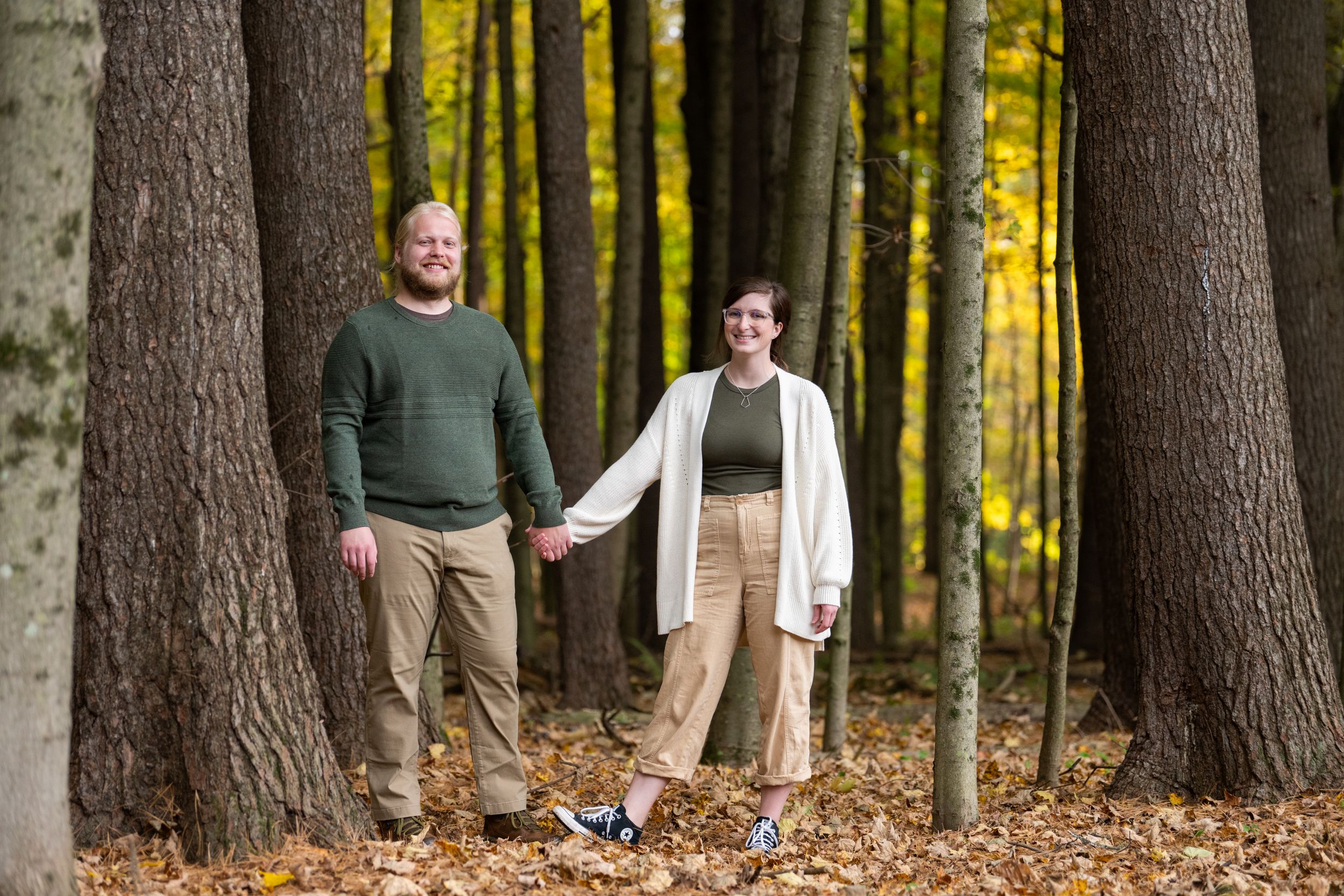 A couple walks hand in hand through a forest of tall trees with autumn leaves on the ground.