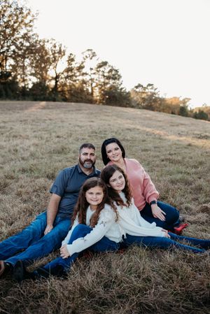 A group relaxes together on a grassy hillside during golden hour as the sun sets behind trees in the distance.