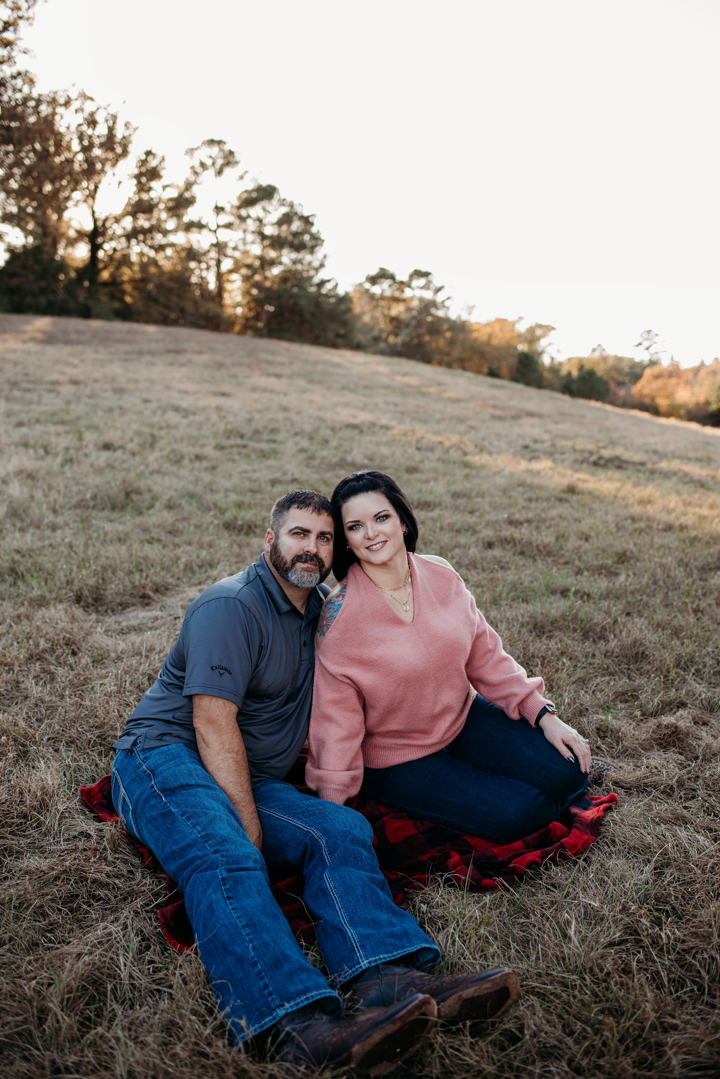 A couple sits together on a grassy hillside during golden hour in a casual outdoor setting.