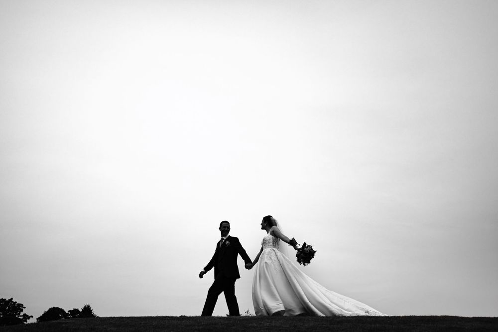 Silhouetted wedding couple and photographer creating artistic black and white portraits against bright sky at sunset.