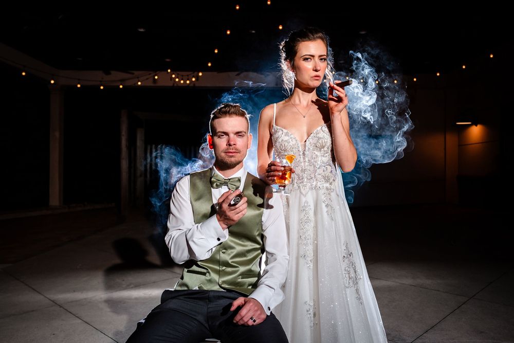Wedding couple creates artistic smoke effect photos in dimly lit venue with string lights in background.