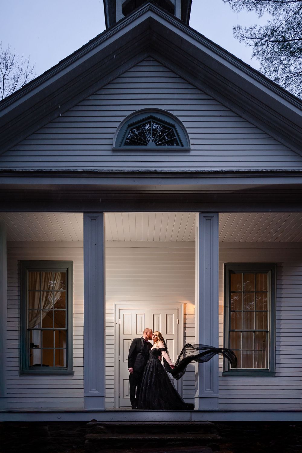Silhouettes visible through windows of a traditional white house with dark trim at dusk.