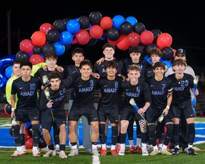 Soccer team in navy uniforms poses together under red and blue balloon arch on sports field at night.