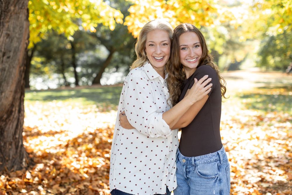 Two smiling women hugging outdoors in a park surrounded by colorful autumn foliage and fallen leaves.