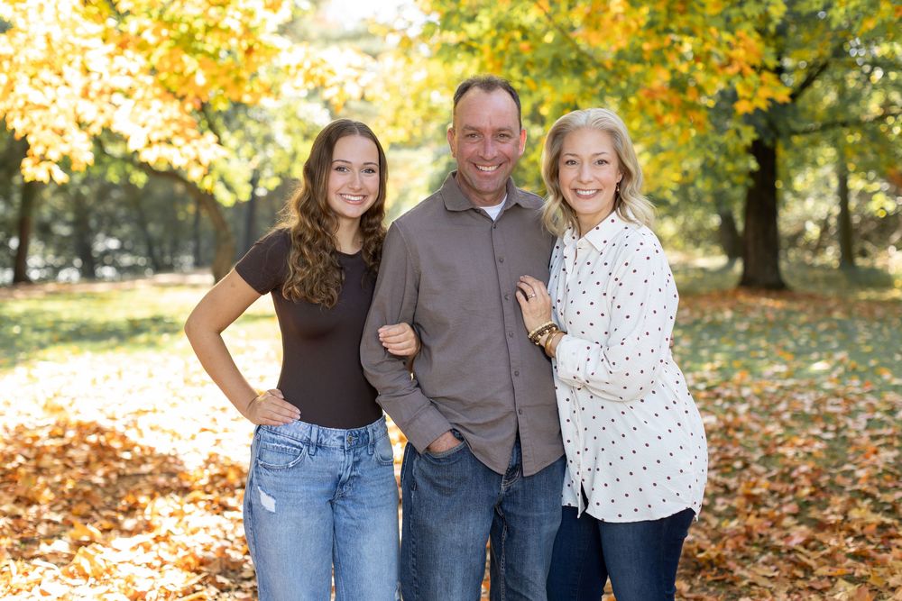 Family of three smiling outdoors in autumn park with colorful yellow and orange foliage in background.