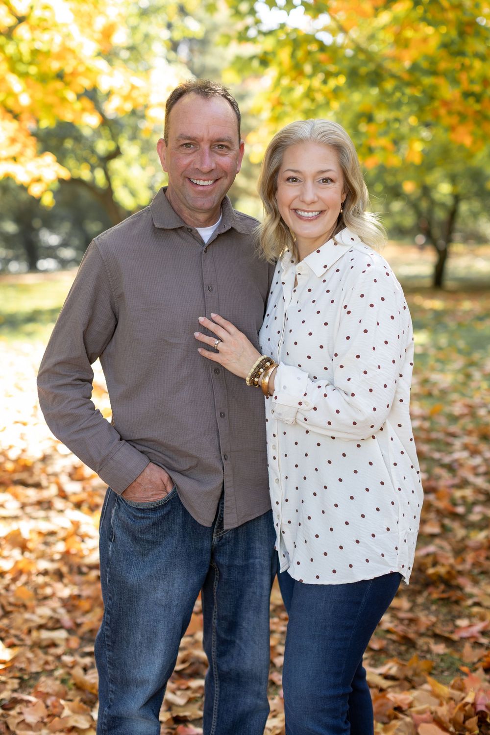 A smiling couple poses together among autumn leaves in a park, wearing casual attire on a sunny fall day.