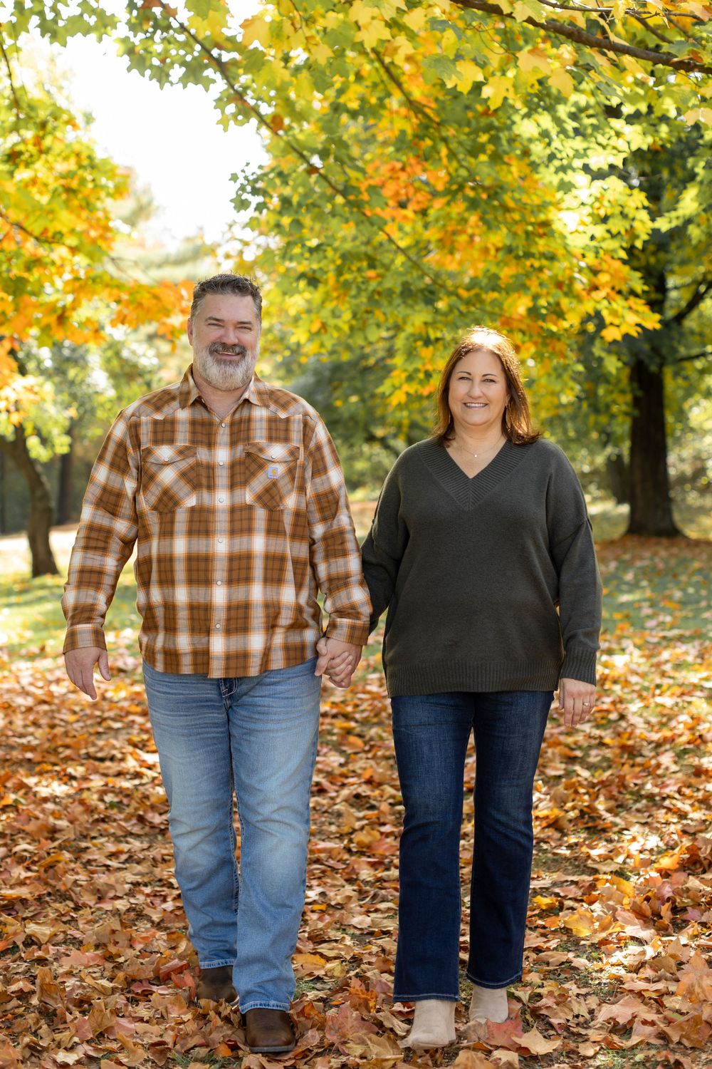 A middle-aged couple holds hands while walking through a colorful autumn park covered in fallen leaves.