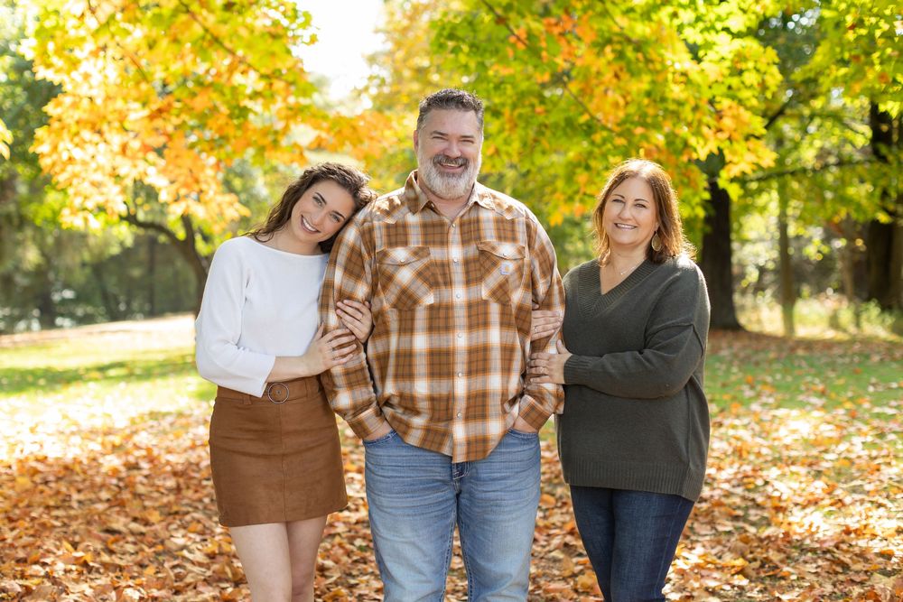 Family of three smiling together in an autumn park surrounded by colorful fall foliage and golden leaves.