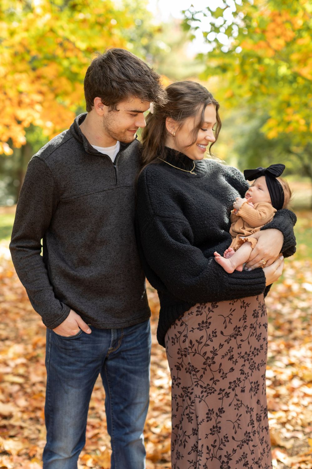 Young couple in dark sweaters admiring their newborn baby in an autumn park with golden foliage.