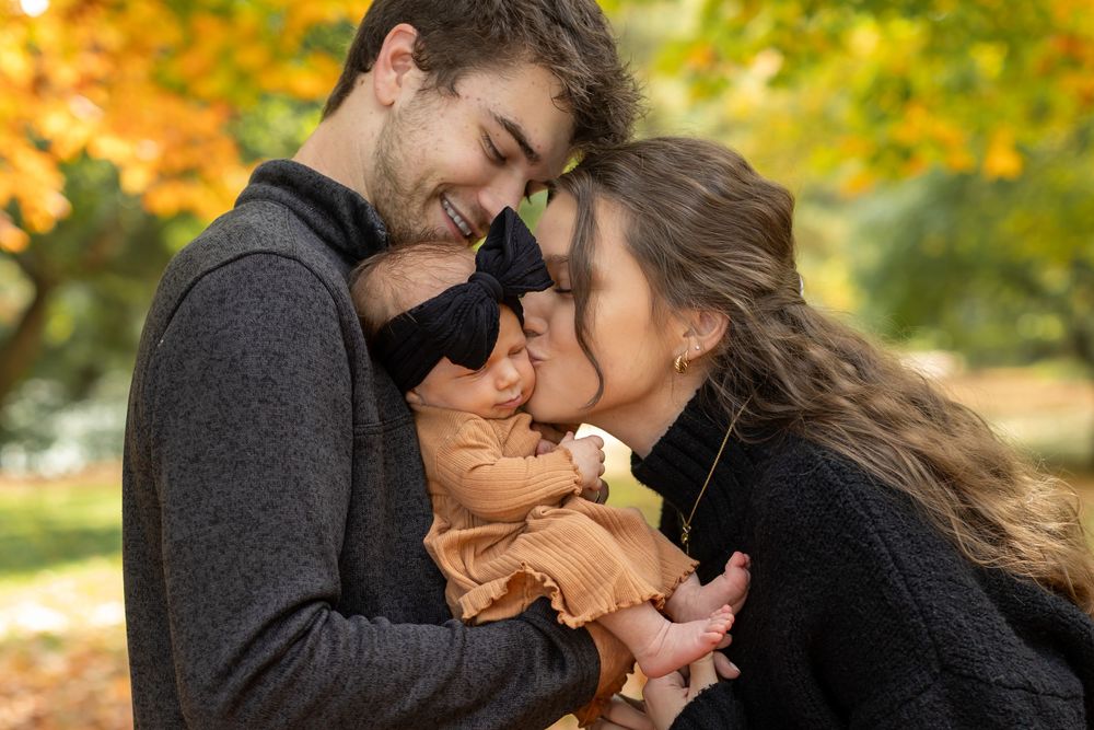 Young couple kissing their newborn baby outdoors in autumn park with colorful fall foliage background.