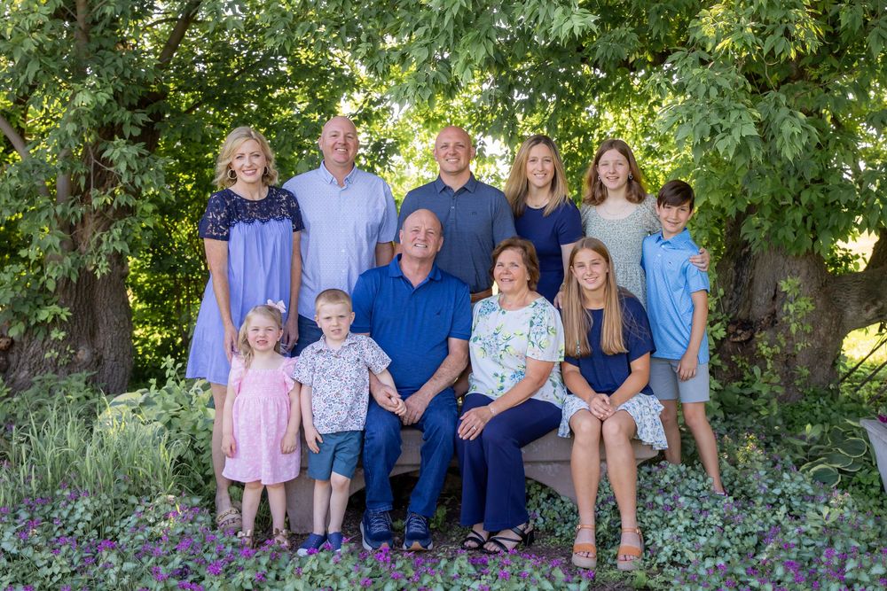 Large family group portrait outdoors in lush green garden setting, three generations smiling together in coordinated blue outfits.