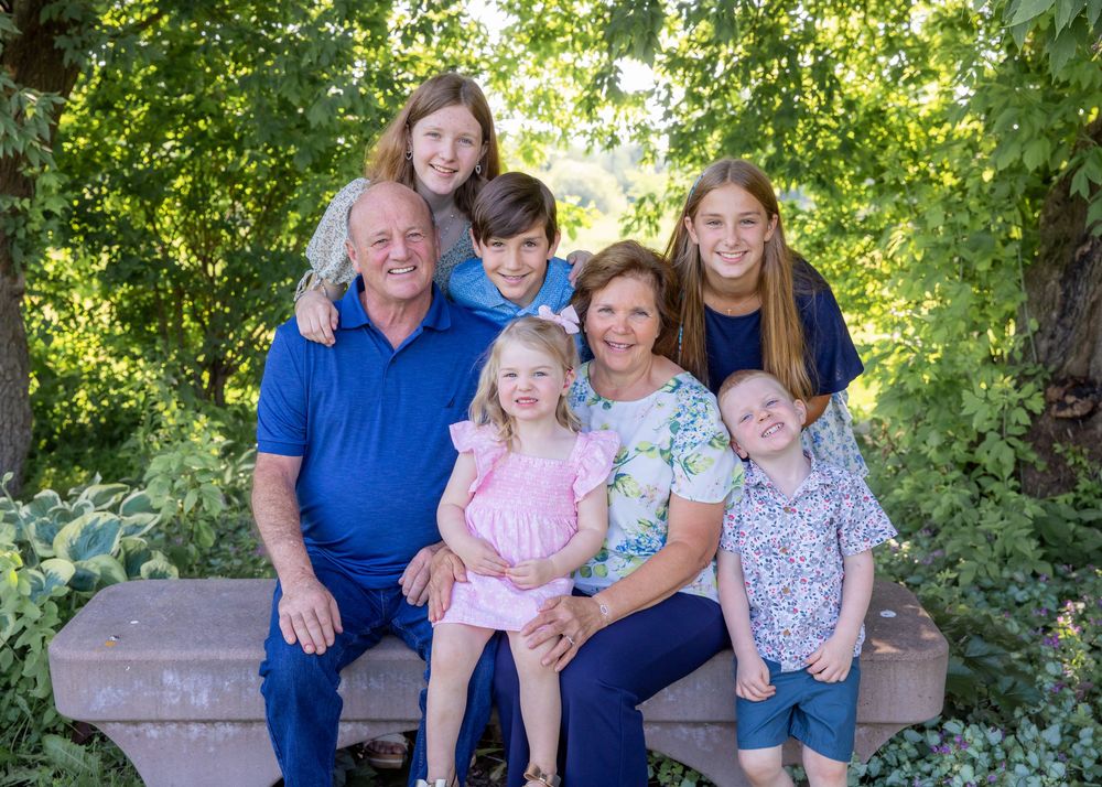 A joyful multigenerational family of seven poses together outdoors on a stone bench surrounded by lush green trees.
