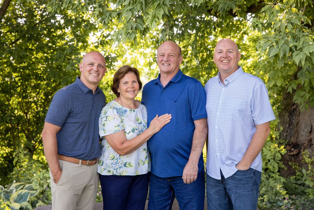 Four smiling adults pose together outdoors surrounded by lush green trees in a warm family portrait.