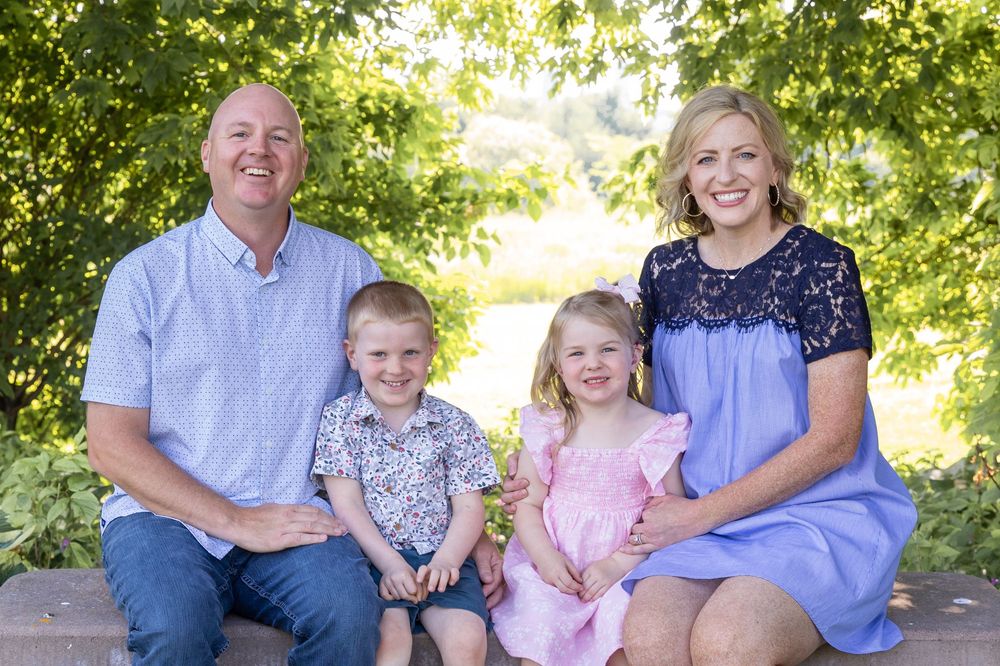 Happy family of four smiling outdoors, parents with young boy and girl in pink dress, surrounded by lush green trees.