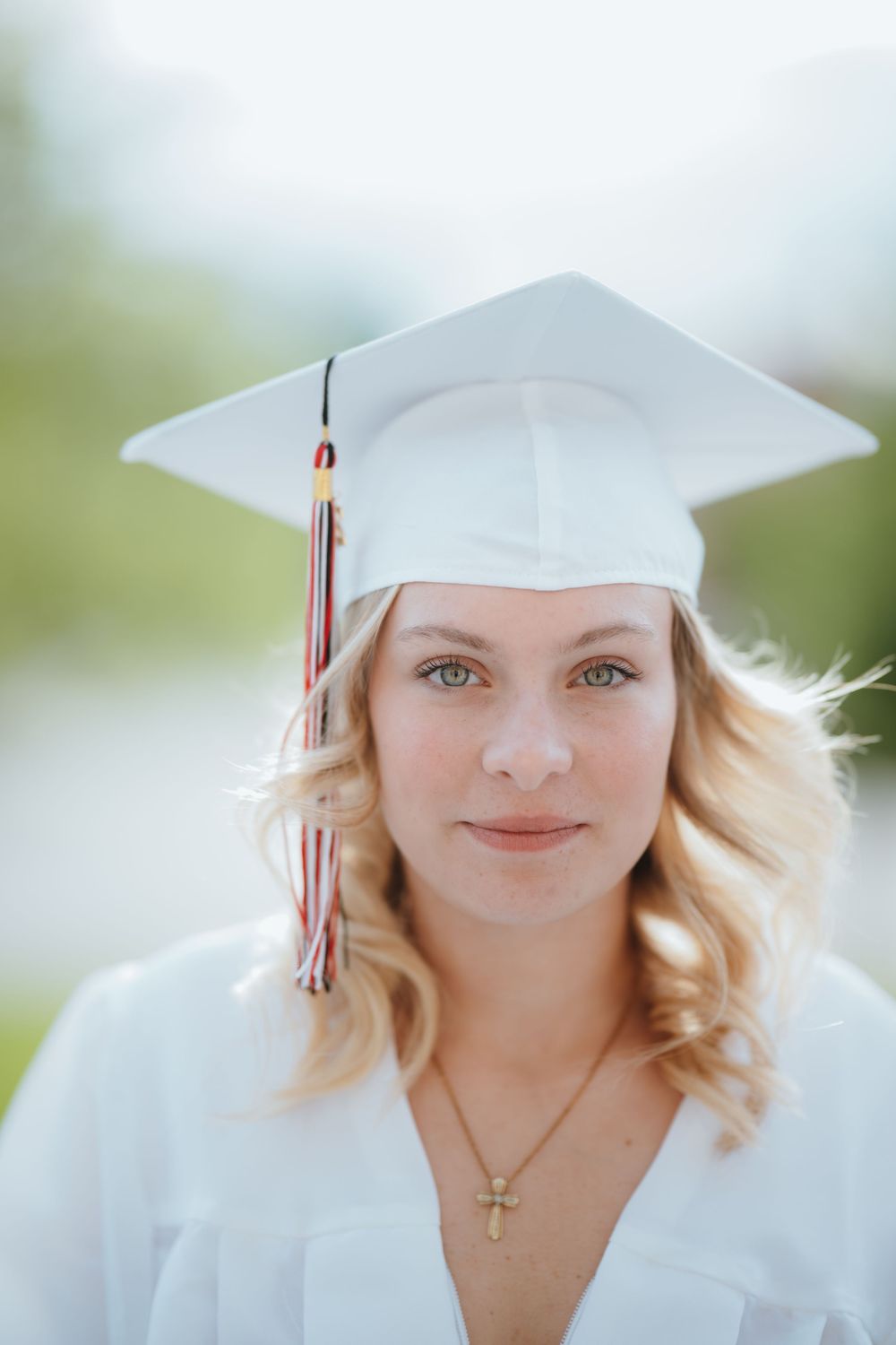 Young woman in white graduation cap and gown smiling, wearing a cross necklace with colorful tassel.