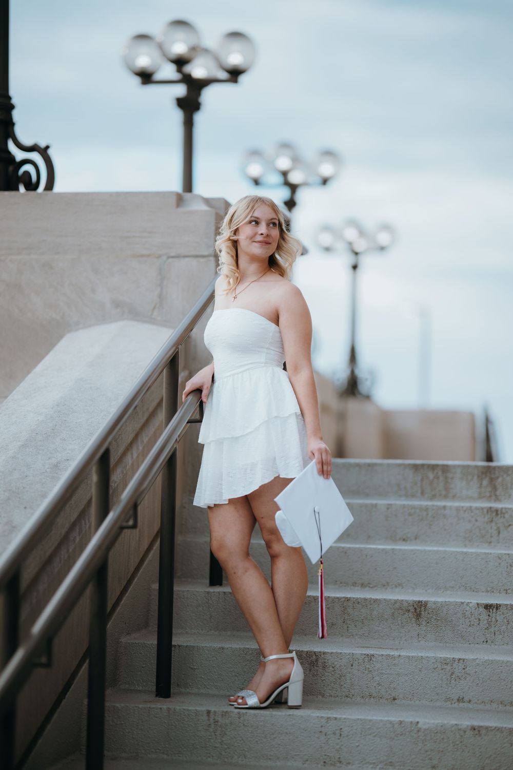 Young woman in white strapless dress holding graduation cap on stone steps with ornate lamp posts behind her.