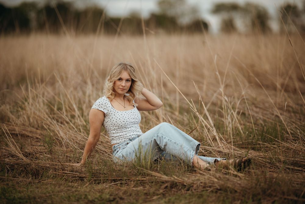 Blonde woman sitting in tall dry grass field wearing white top and light blue jeans in outdoor portrait photo.