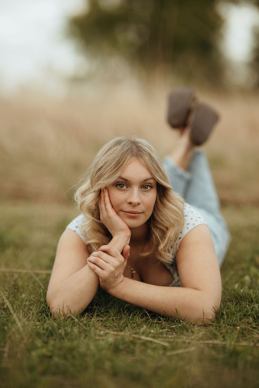 Blonde woman lying on grass in a light blue outfit, resting her chin on her hand, smiling softly outdoors.