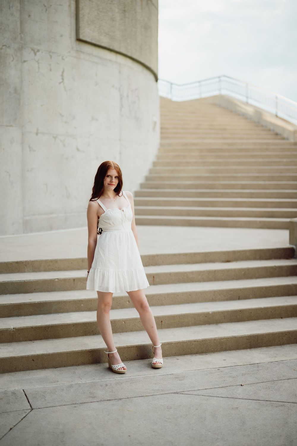 Young woman in white sundress and heels poses gracefully on concrete steps near curved architectural wall.