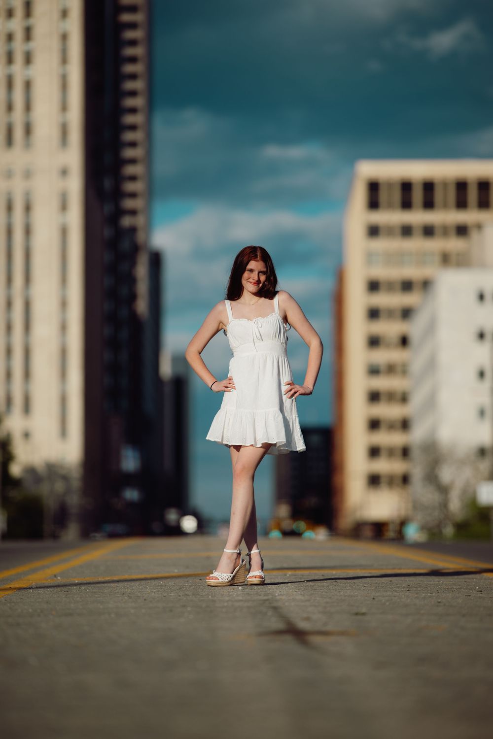 Young woman in white dress poses confidently on urban street with city buildings in background on sunny day.