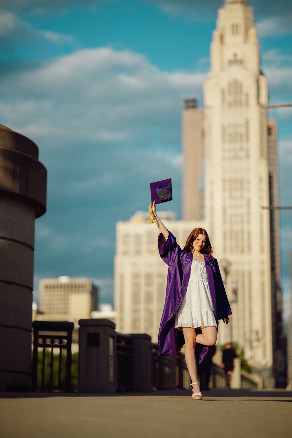 Young woman in purple graduation gown raises cap triumphantly on urban bridge with city skyline and skyscraper behind her.