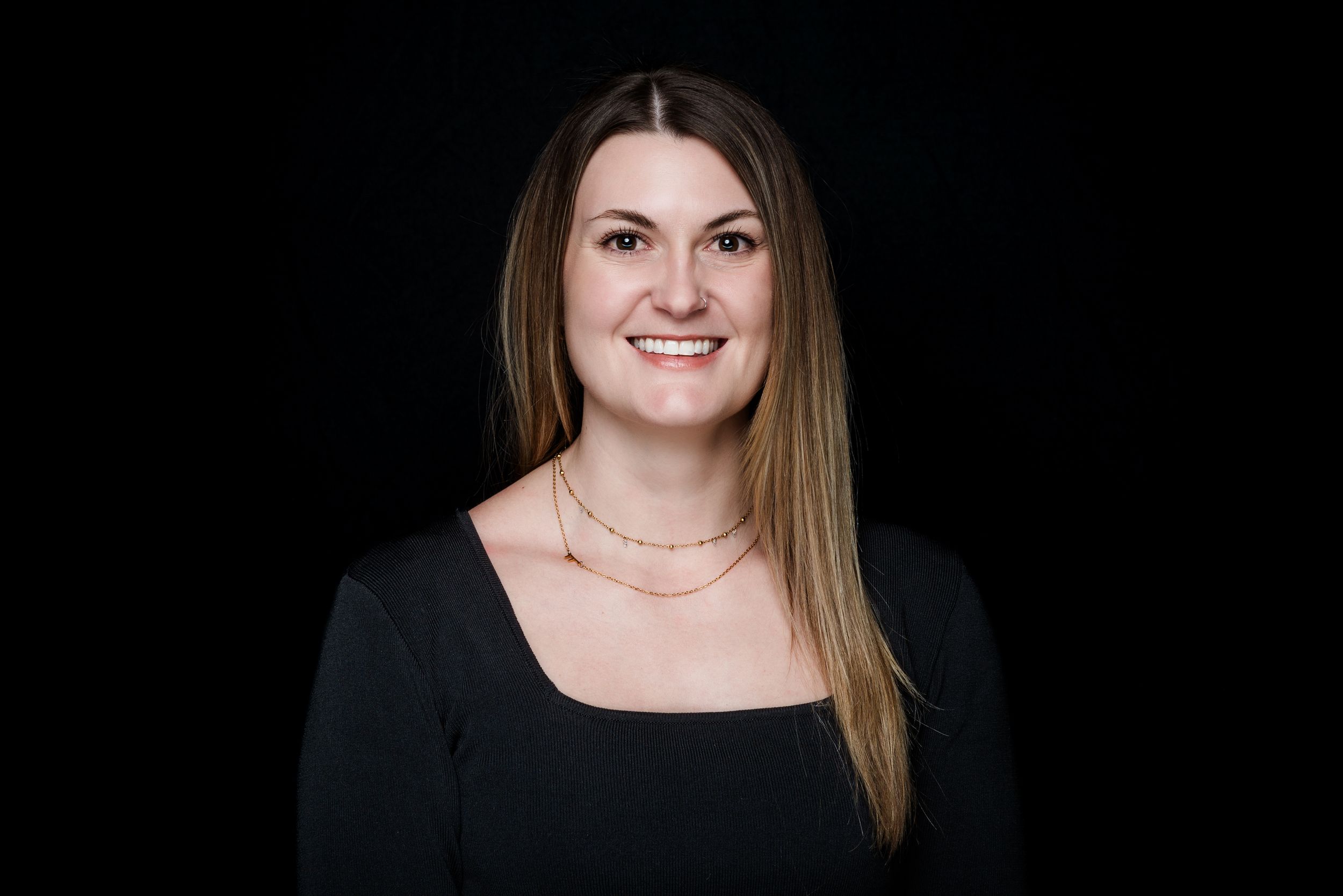 Professional headshot portrait on black background featuring long straight hair, black top and delicate necklace.