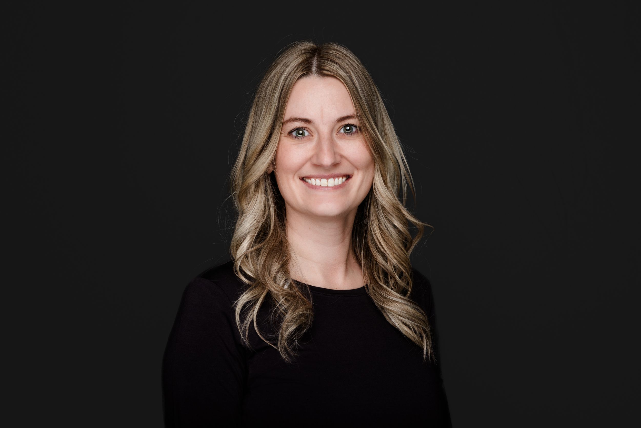 Professional headshot of someone with long blonde wavy hair wearing a black top against a dark background.
