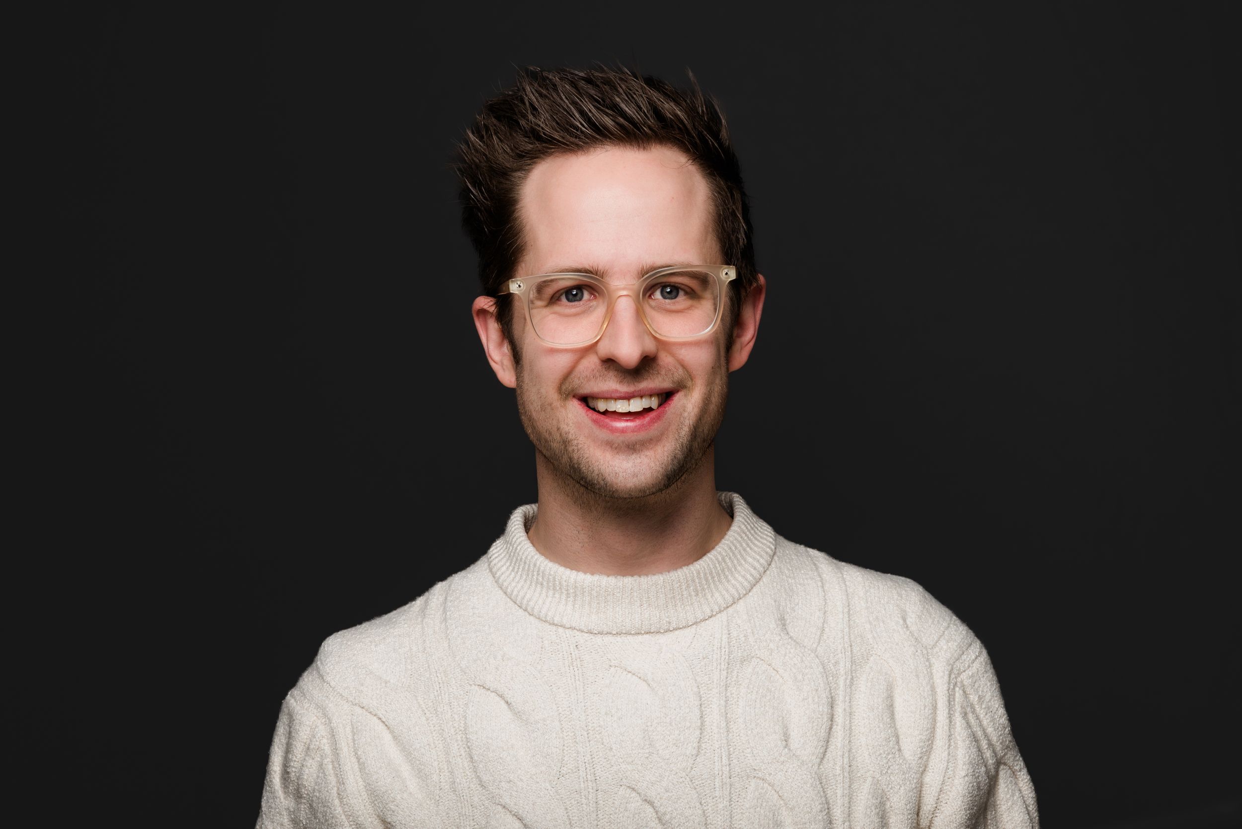 Person wearing white cable knit sweater smiles while posing against dark background.