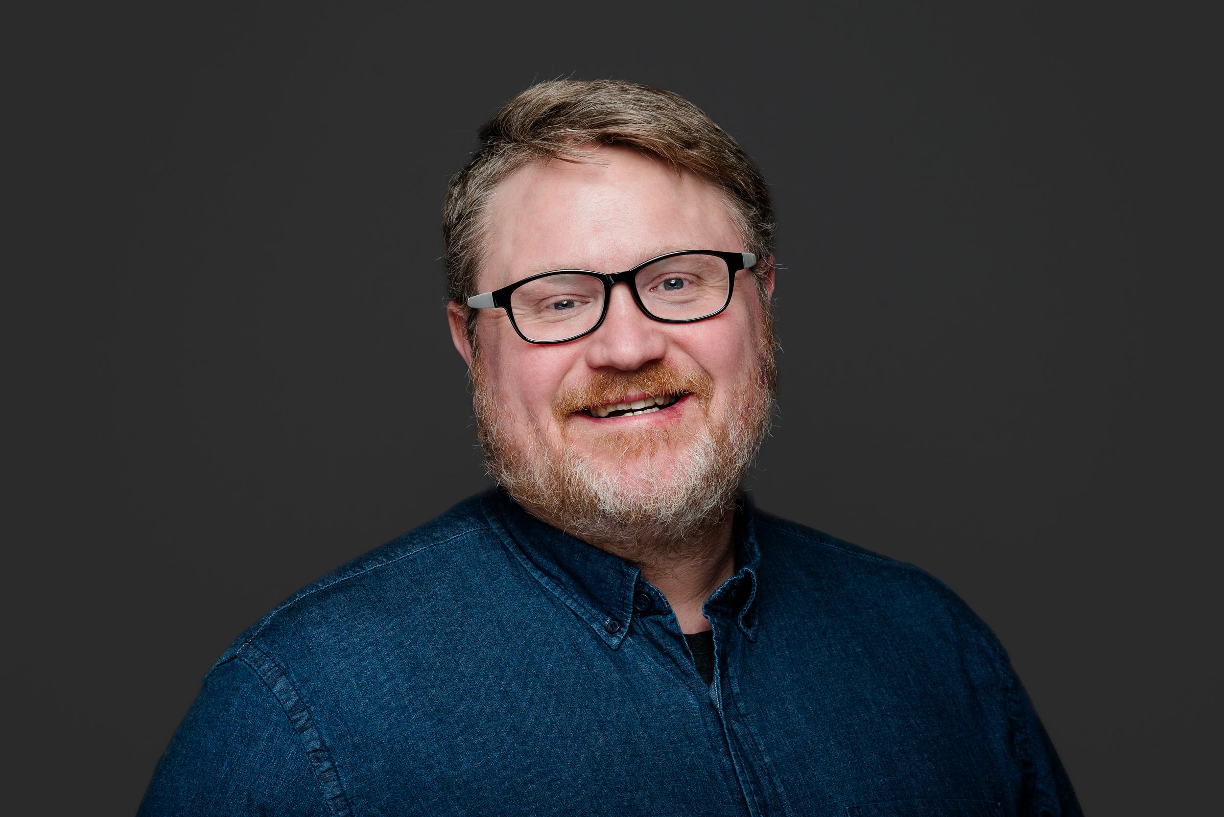 A person wearing glasses and a blue shirt smiles warmly at the camera against a dark background.