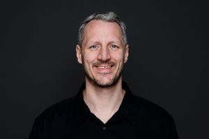 Smiling person wearing a black v-neck shirt against dark background.