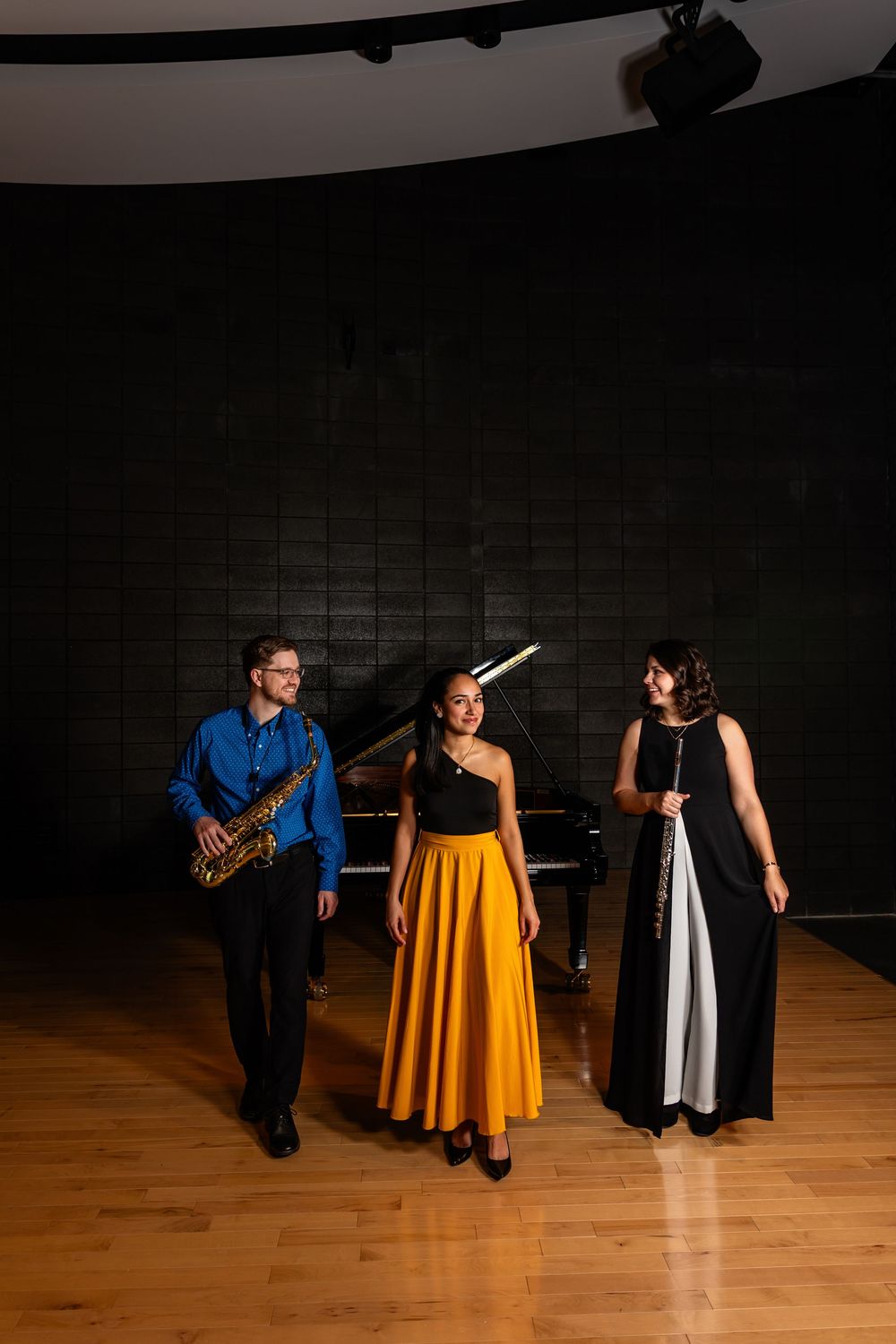 Three musicians in formal attire perform with saxophone, piano and flute on a dark stage with wooden floors.