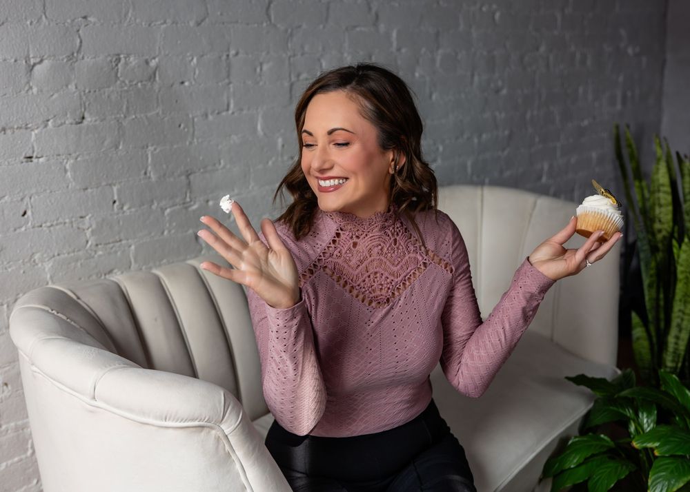 Person in pink sweater sits in grey chair holding cupcake with joyful expression.