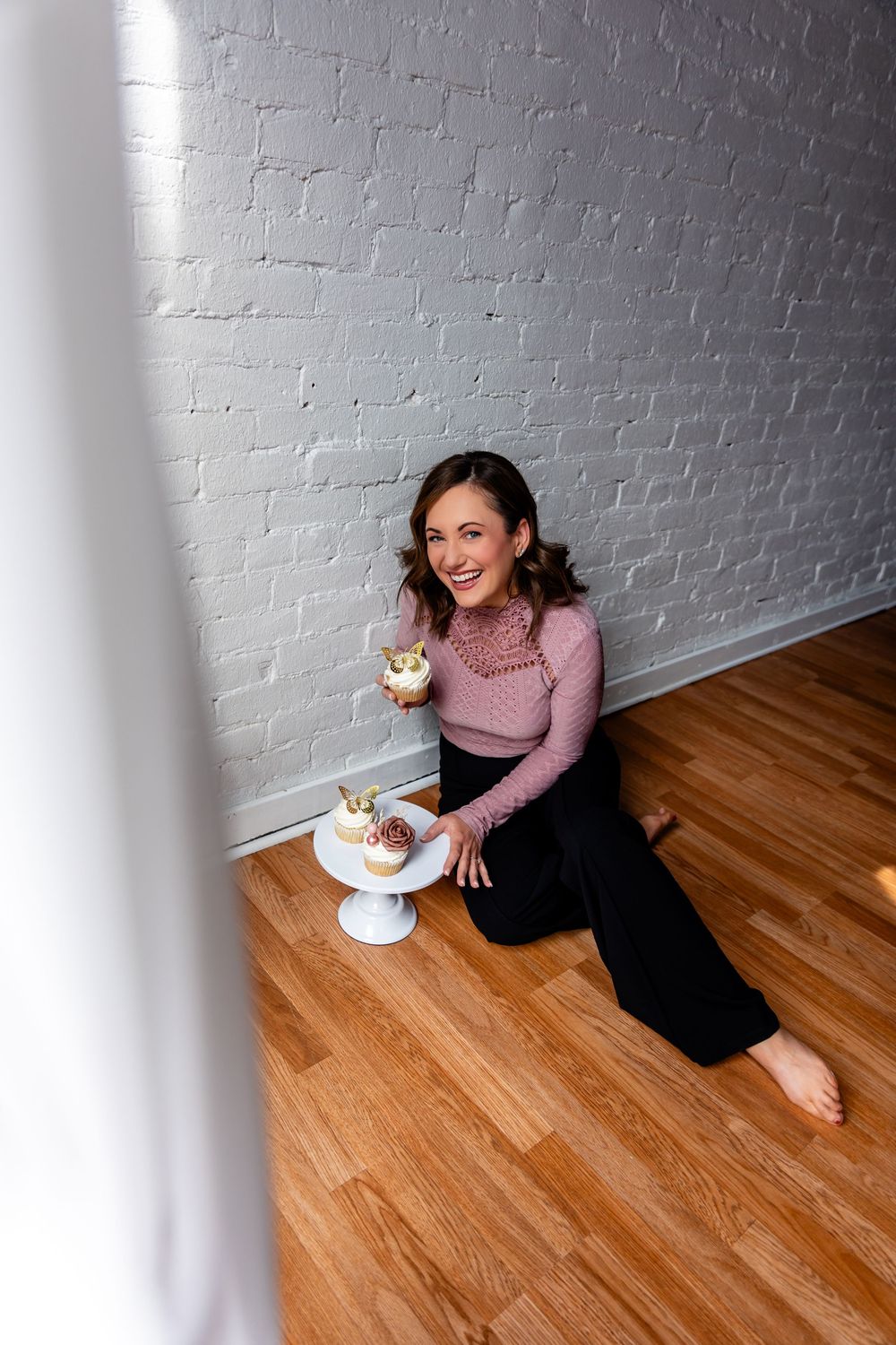 Person in pink sweater sits on wooden floor enjoying cupcake near white brick wall.