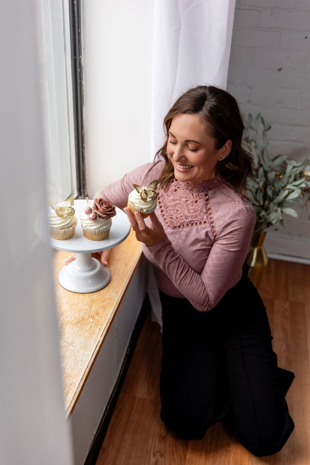 Person in a pink sweater enjoying a cupcake by a window in a cozy cafe setting.