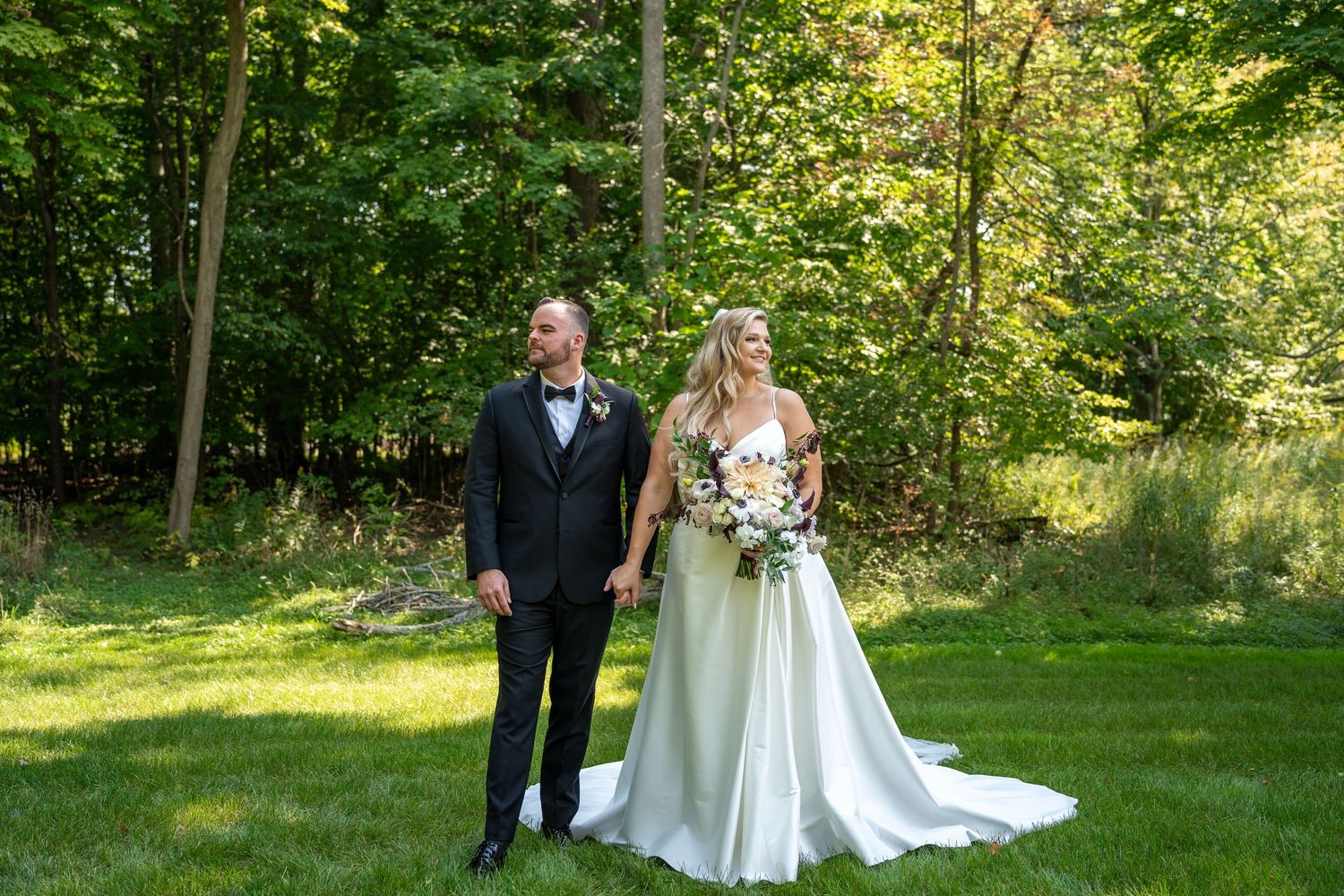 A couple in wedding attire holding hands while standing on a lush green lawn surrounded by trees on a sunny day.