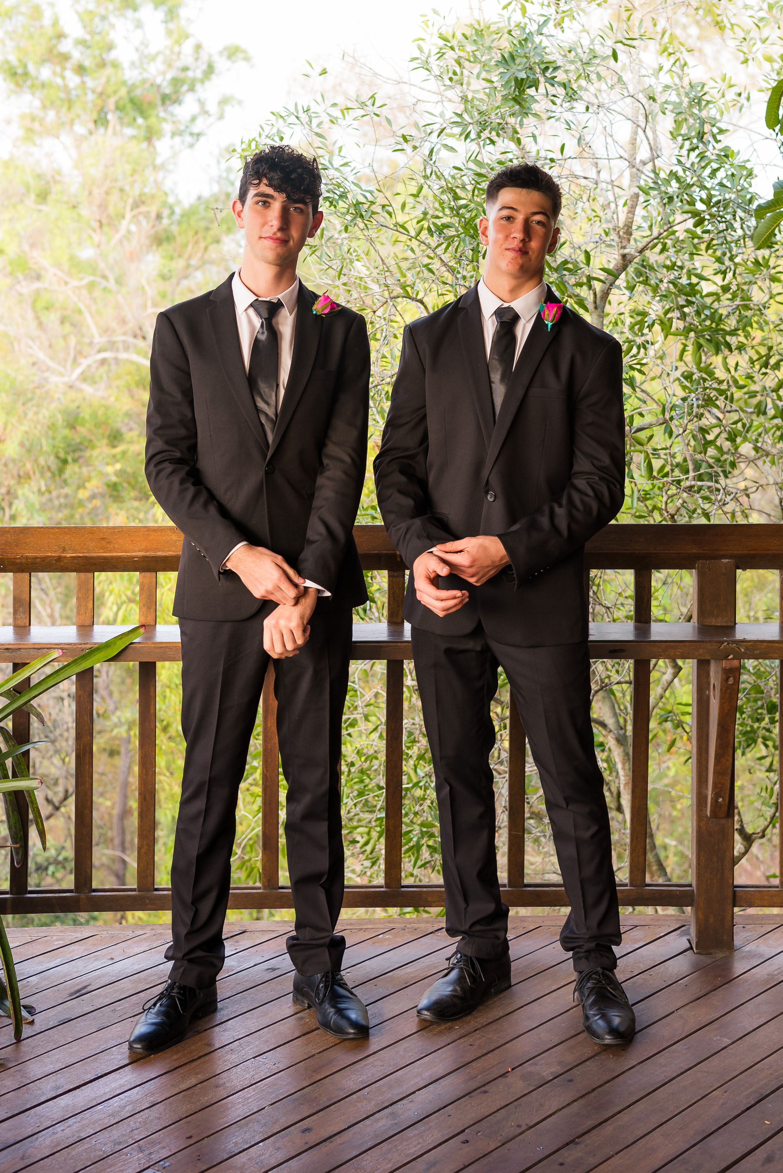 Four people wearing black suits stand in a row on a wooden deck with greenery visible in the background.