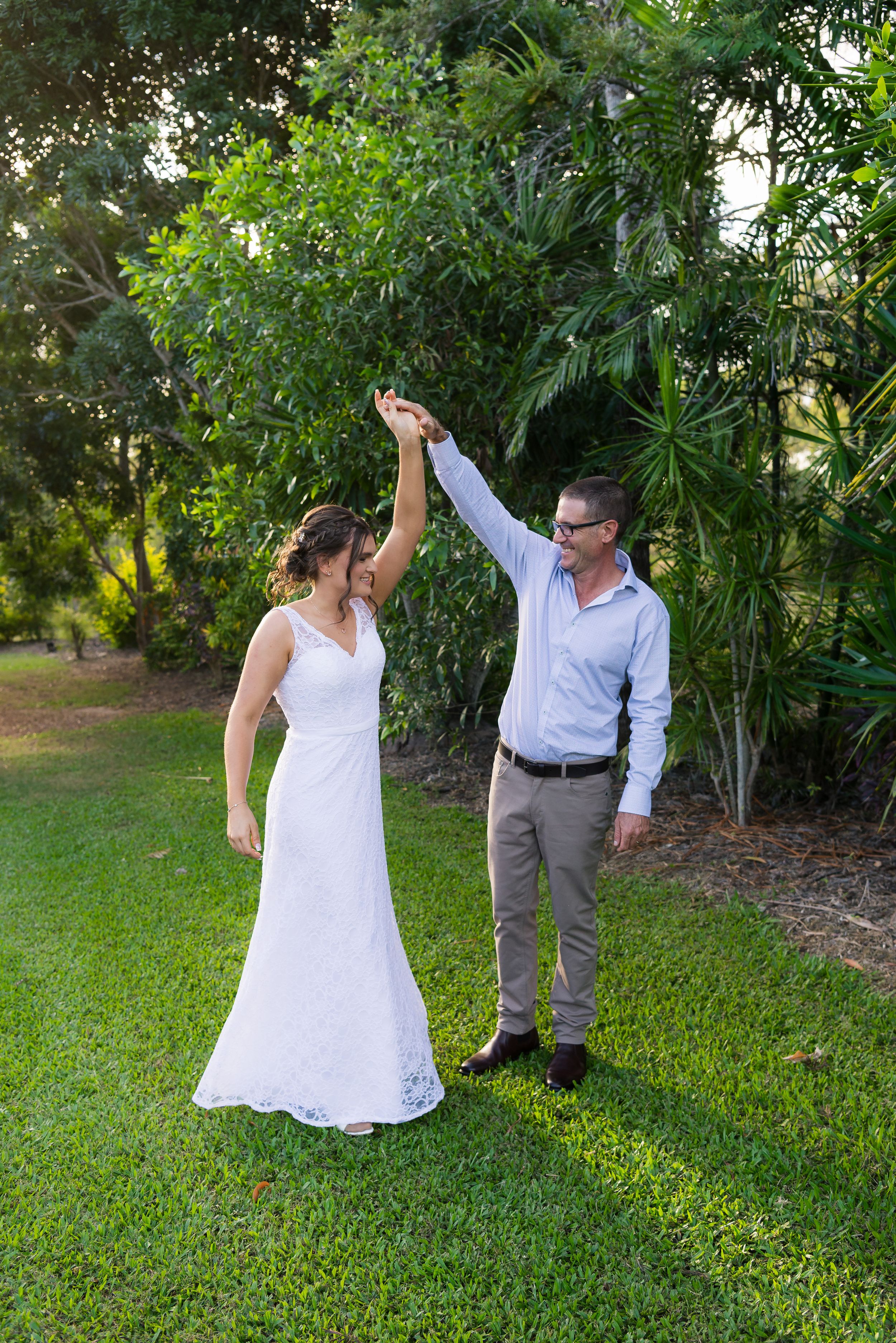 A couple dances together on a lush green lawn surrounded by tropical plants during a casual outdoor celebration.
