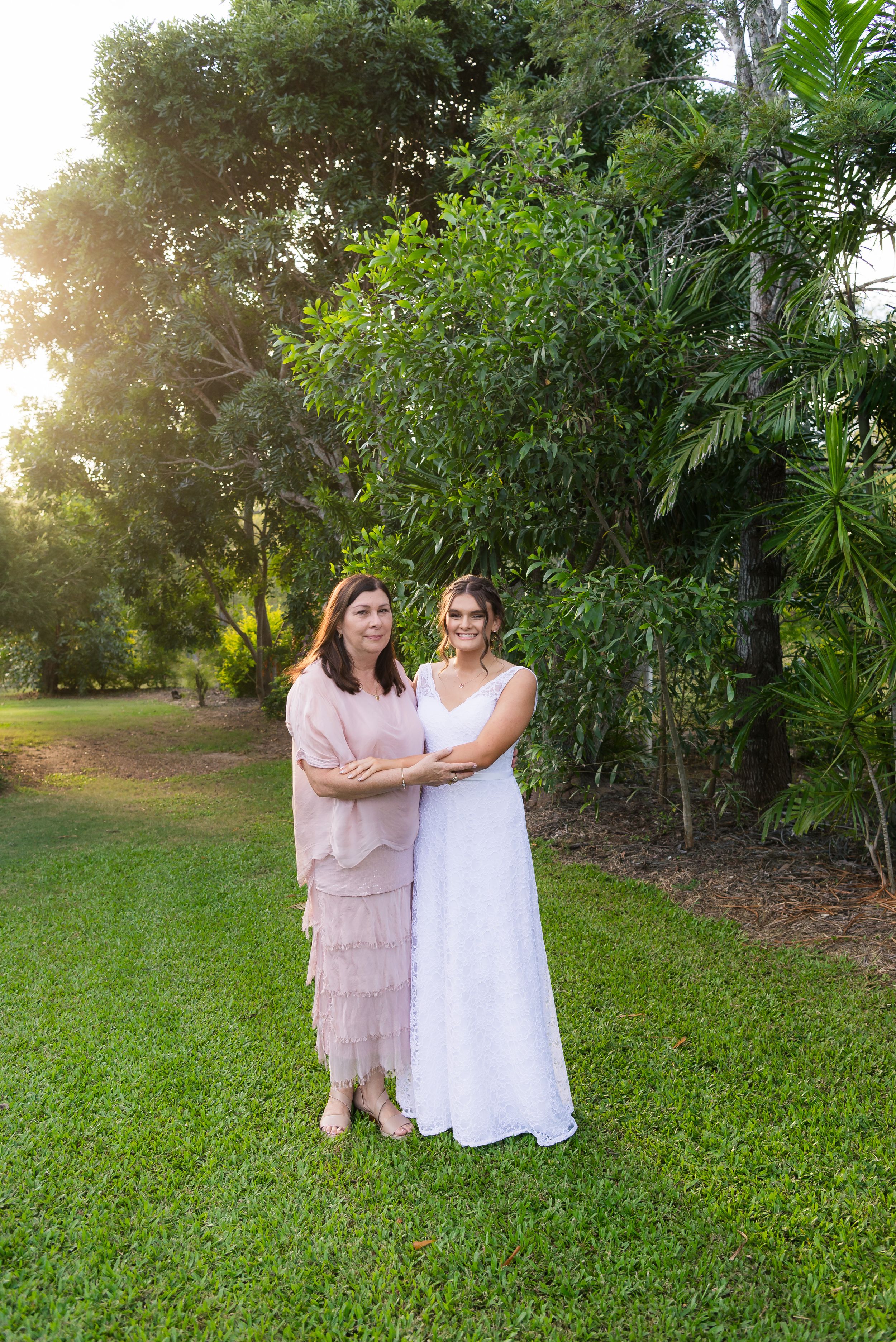 Two people in light colored dresses stand together on green grass near tropical foliage at sunset.