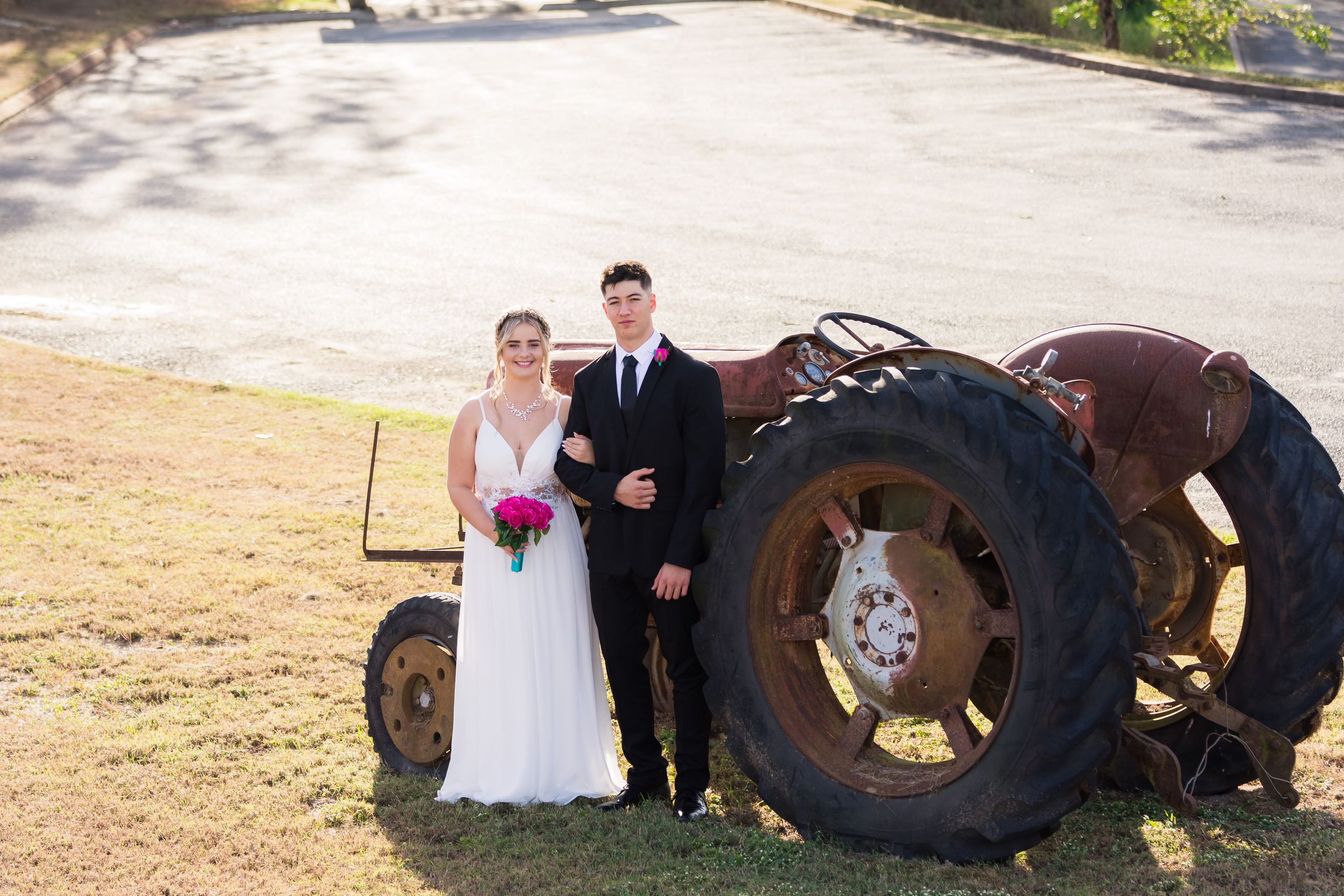 A rustic outdoor wedding photo with large vintage tractor wheels and bright pink bouquet.