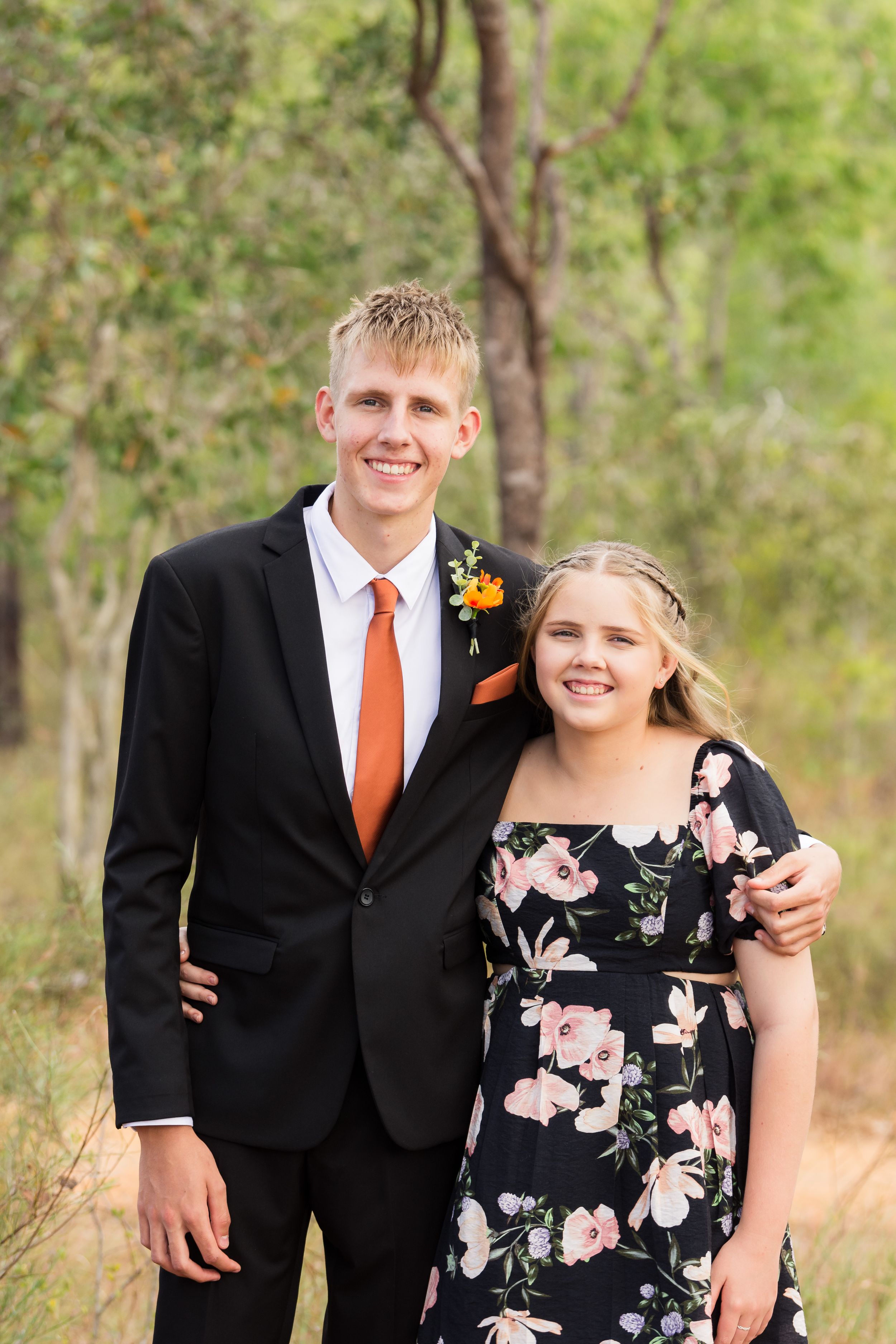 A couple poses outdoors in formalwear against a natural backdrop on a sunny day.